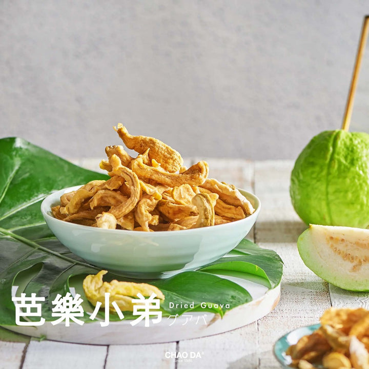 Dried guava slices in a white bowl with green leaves and guava fruit on a light wooden surface.