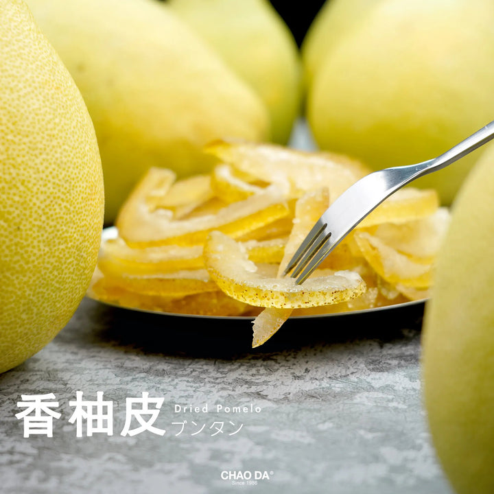 Dried pomelo slices on a plate with a fork, surrounded by whole pomelos, on a gray surface.